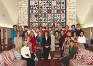 Photo of Reba McEntire and the staff at Reba's Ranch House posing for a photo infront of a large quilt in the main lounge area of Rebas Ranch House